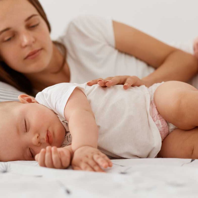 young-adult-female-with-dark-hair-lying-with-baby-bed-looking-daughter-sees-sleeping-she-woman-wearing-white-casual-t-shirt-happy-motherhood